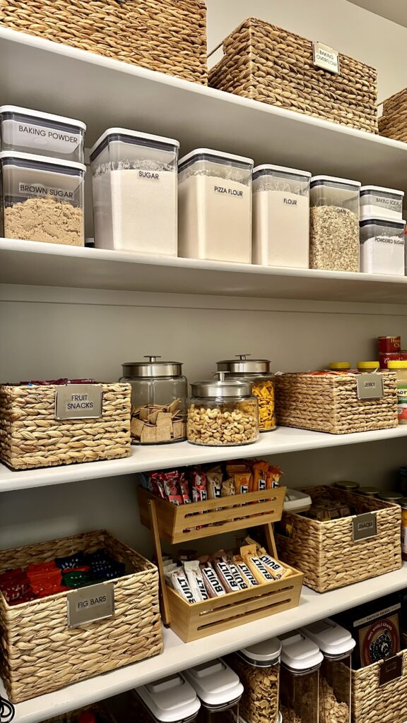 Organized pantry with labeled baskets, clear containers of dry goods, and jars of snacks arranged neatly on white shelves