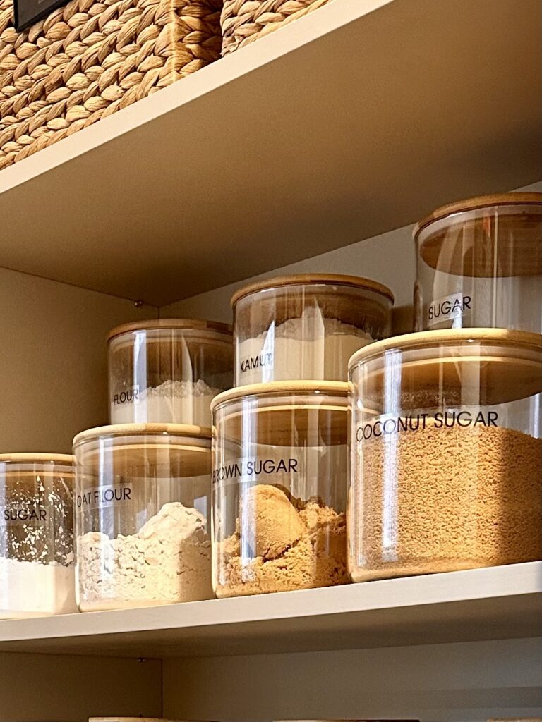 Glass pantry jars labeled with sugars and flours, including coconut sugar, brown sugar, oat flour, and kamut, neatly arranged on a shelf