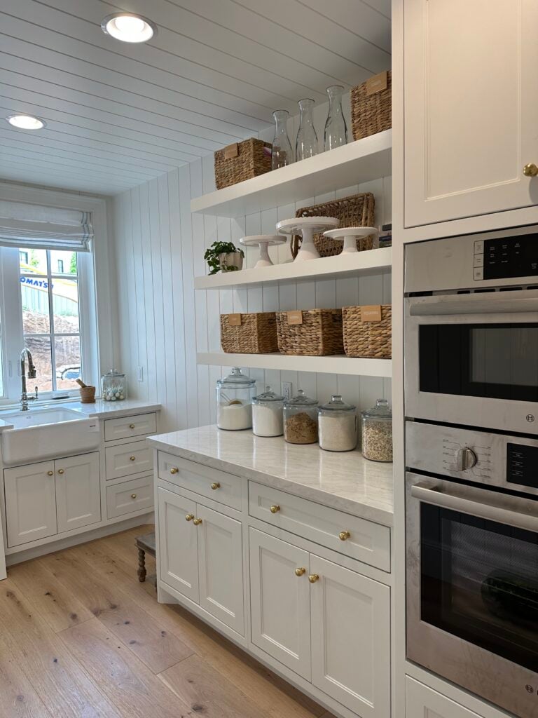 Bright white kitchen with open shelving, labeled woven baskets, glass canisters of pantry staples, and double wall ovens