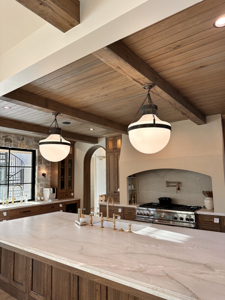 Luxury kitchen with wood beams, large marble island, globe pendant lights, brass fixtures, and built-in range under arched hood