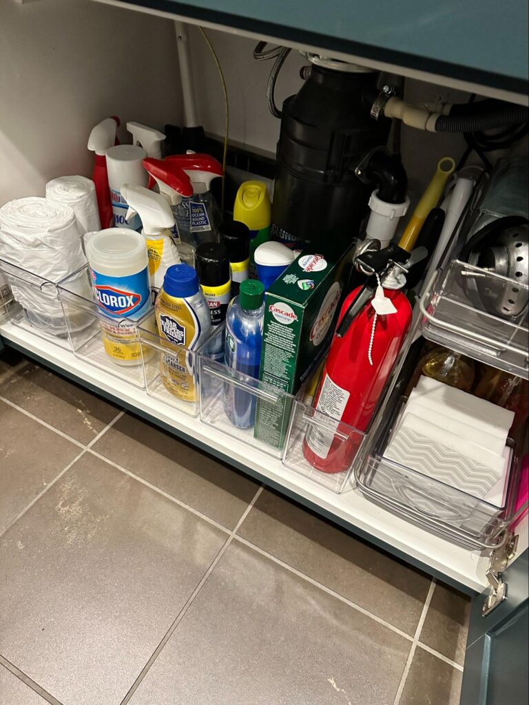 Organized cleaning supplies neatly stored under a kitchen sink with sprays, wipes, and paper towels.