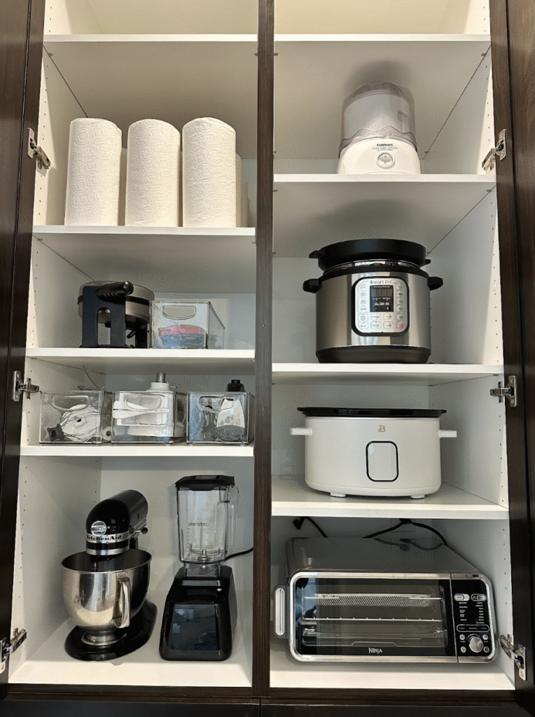 Tall cabinet organized with kitchen appliances, paper towels, and clear bins storing accessories.
