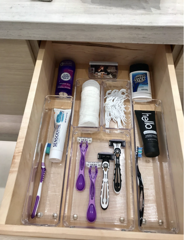 Bathroom drawer organized with clear trays holding razors, toiletries, cotton pads, flossers, and toothpaste.