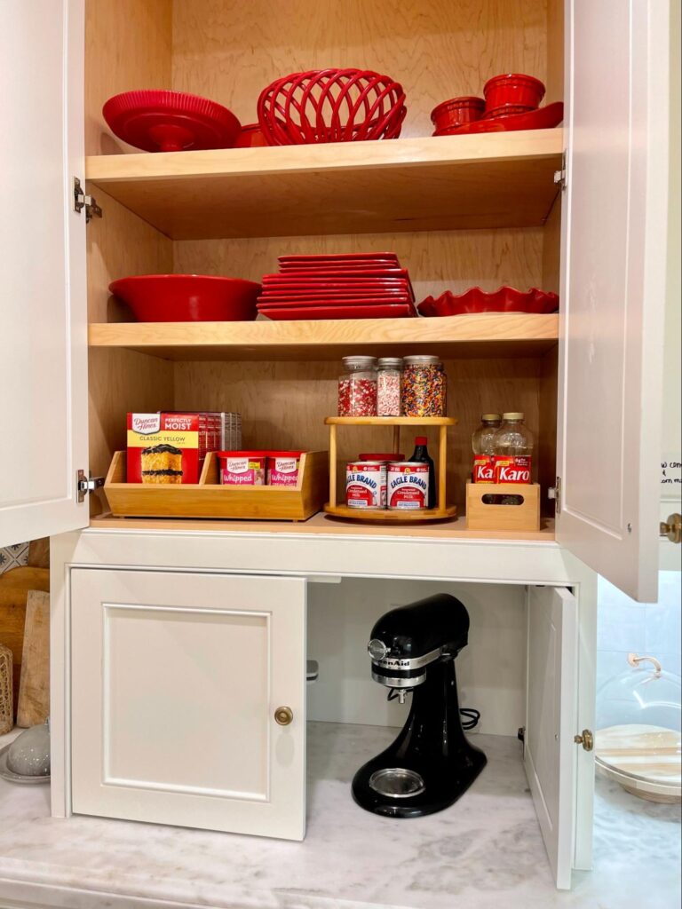 Cabinet stocked with red holiday dishes and baking supplies, with jars of sprinkles and a mixer stored below.