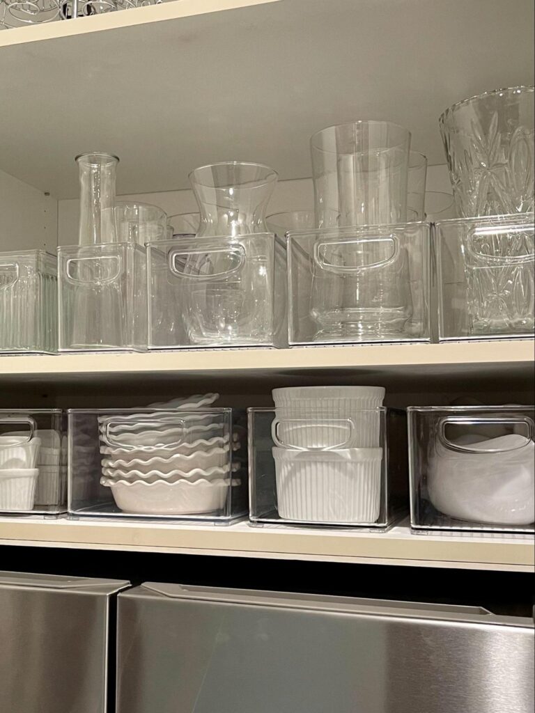 Clear bins holding glass vases and white dishes arranged neatly on open shelves.