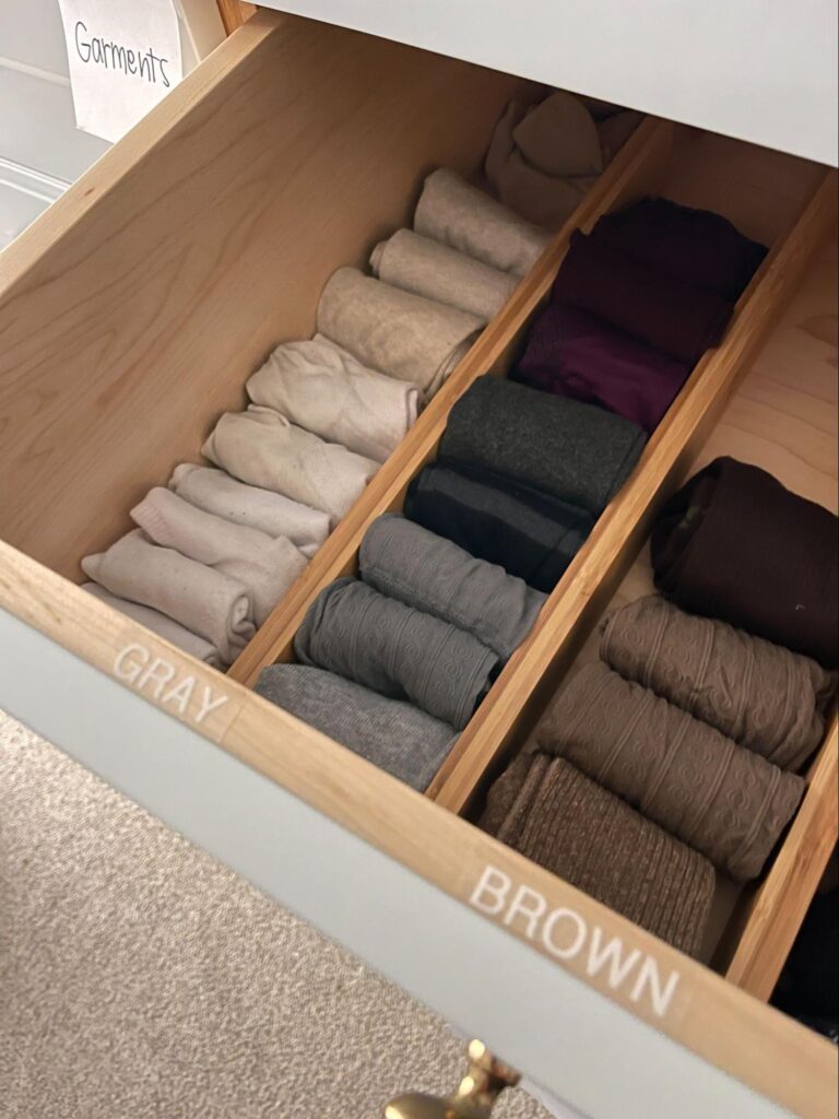 Organized drawer with folded clothing sorted by color using wooden dividers.