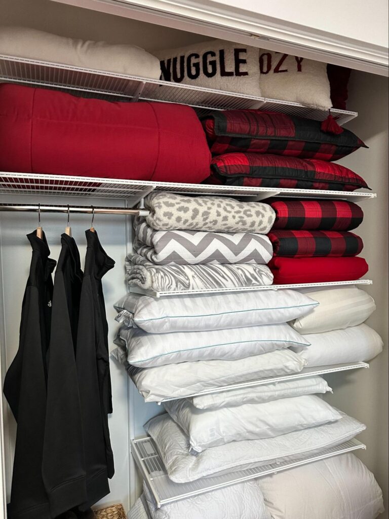 Closet shelves with neatly folded blankets, pillows, and hanging black clothing.
