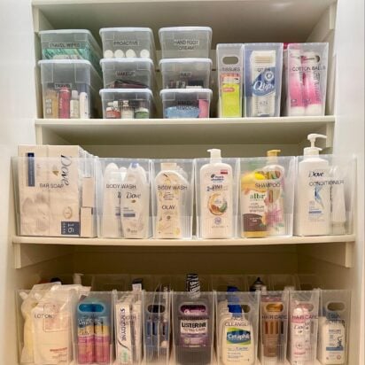 Organized linen closet with clear bins holding toiletries, soaps, hair products, and personal care items on labeled shelves