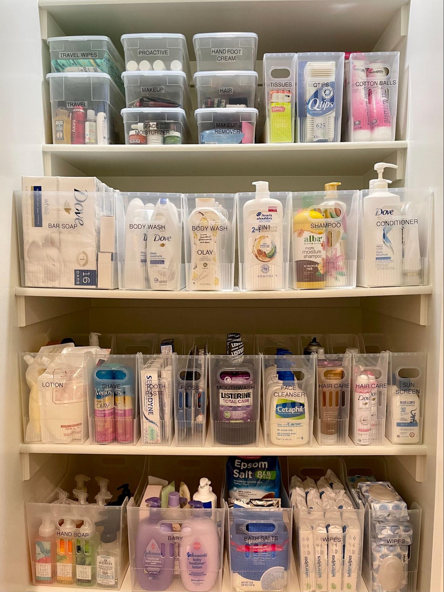Organized linen closet with clear bins holding toiletries, soaps, hair products, and personal care items on labeled shelves