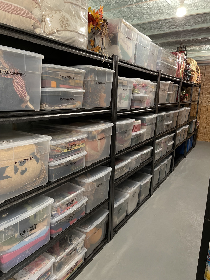 Organized basement storage room with black metal shelving and labeled clear plastic bins for Halloween, Thanksgiving, and Christmas decorations.