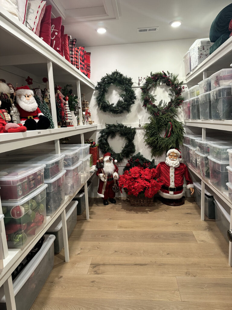 Walk-in storage room with clear plastic bins on white shelving and Christmas decorations arranged along the walls.