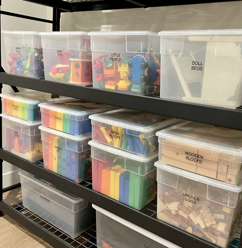 Clear plastic storage bins filled with colorful craft supplies arranged neatly on black metal basement shelves.